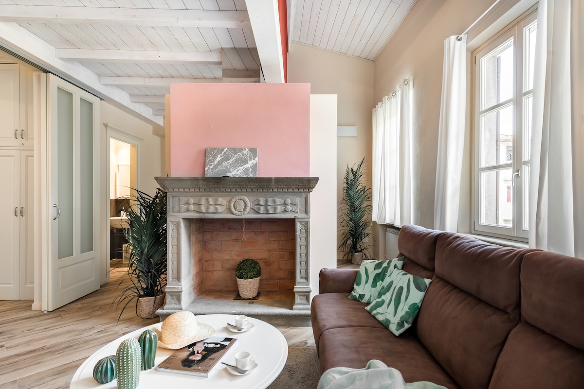 A spacious living area features a welcoming brown sofa arranged around a circular white coffee table, accented by decorative plants. Light enters through large windows, paired with sheer curtains, creating a bright atmosphere. A historic fireplace with a marble mantel adds character to the room.