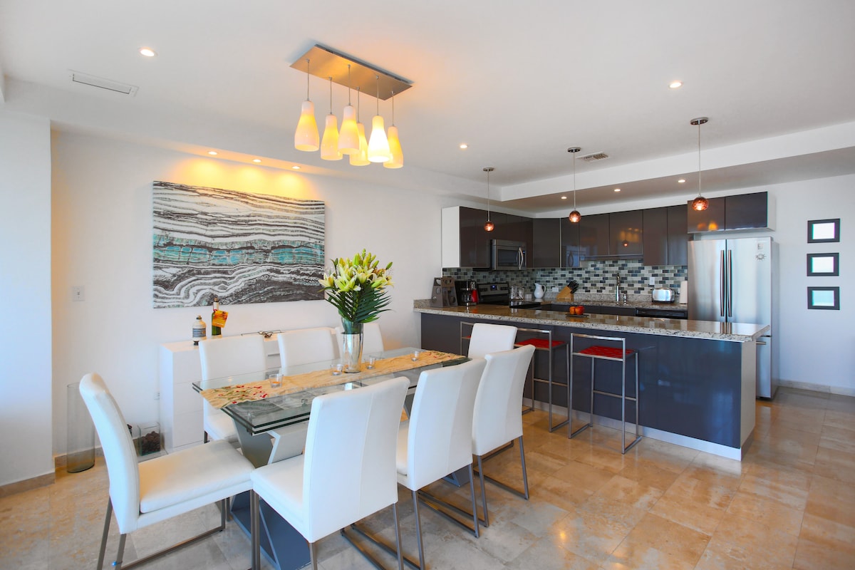 A modern kitchen and dining area is showcased, featuring a sleek dining table with white chairs and a stylish backsplash. The kitchen is equipped with stainless steel appliances and offers ample counter space. Soft pendant lighting illuminates the area, creating a bright and inviting atmosphere.