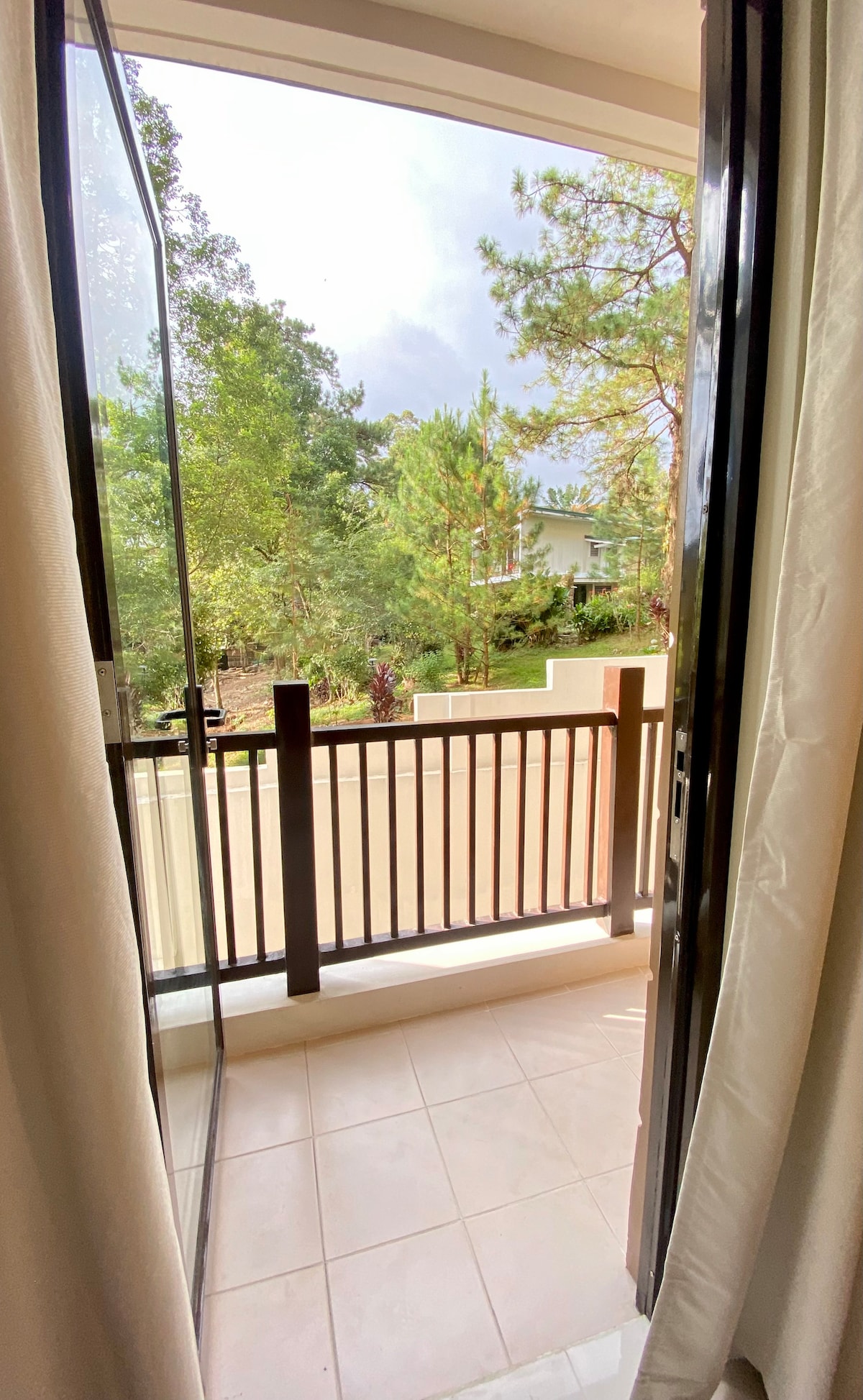 A balcony view is captured, framed by open glass doors. Lush greenery, including pine trees, is visible beyond the railing, offering a serene outdoor space. The tiled balcony floor reflects natural light, enhancing the inviting ambiance of the area.