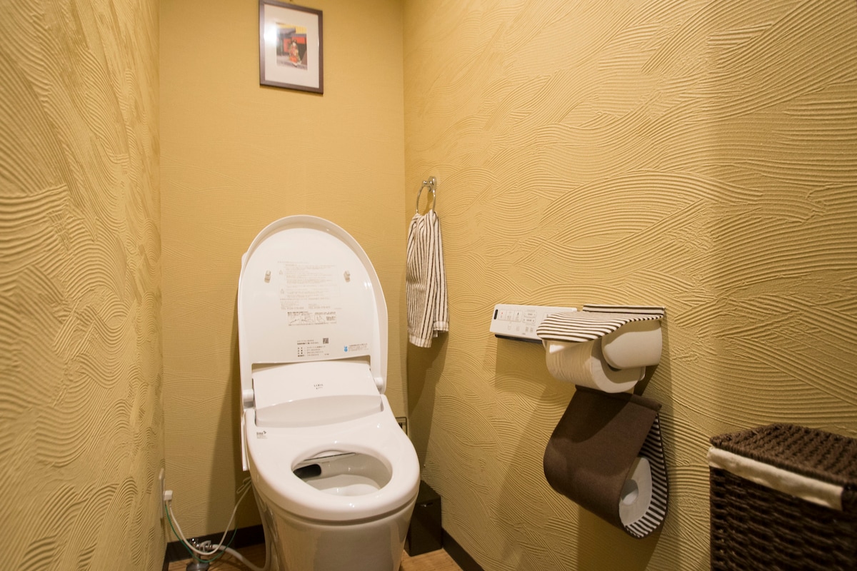 A bathroom featuring a modern toilet equipped with a bidet function, set against textured, warm-toned walls. A framed picture is mounted on the wall, complemented by a towel rack and a toilet paper holder nearby. A woven basket adds a functional touch to the space.