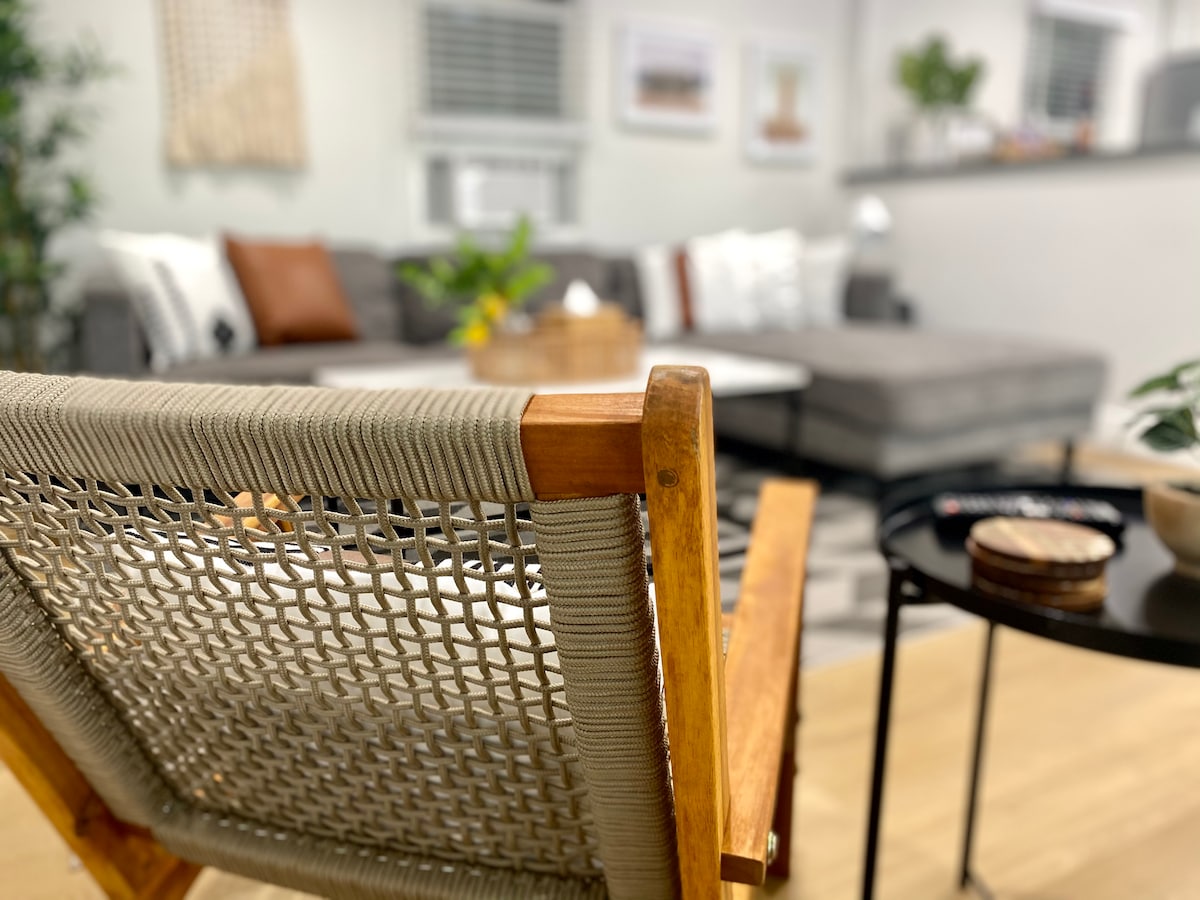 A wooden chair with woven detail is positioned in the foreground, offering a view of the cozy living area. The spacious sectional sofa is adorned with cushions, and a coffee table sits in the center, surrounded by plants and decorative items.