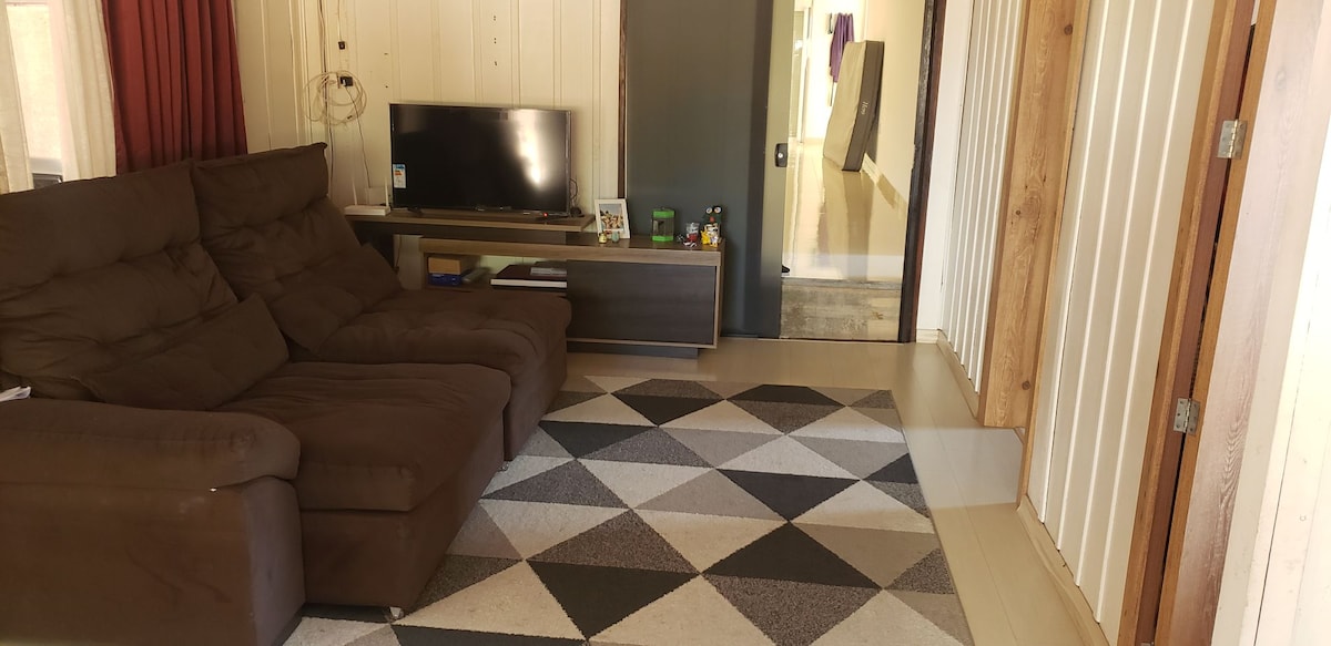 A spacious living area is highlighted by a large brown sectional sofa facing a flat-screen television. A geometric-patterned area rug covers the floor, while natural light is welcomed through the open door leading to a hallway.