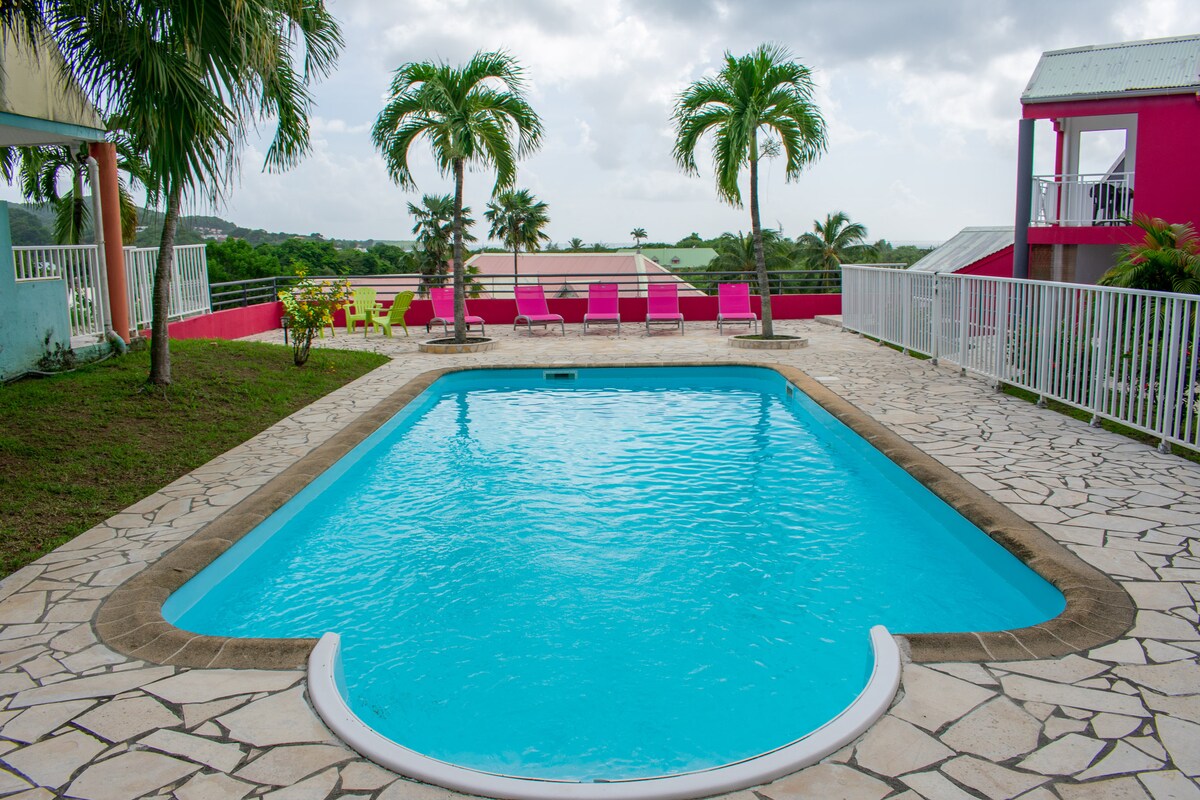 A refreshing pool is framed by a stone patio, with palm trees providing a natural backdrop. Vibrant pink lounge chairs are positioned nearby, and the surrounding area features well-maintained grass. The scene is brightened by soft natural light and an expansive view of the landscape.