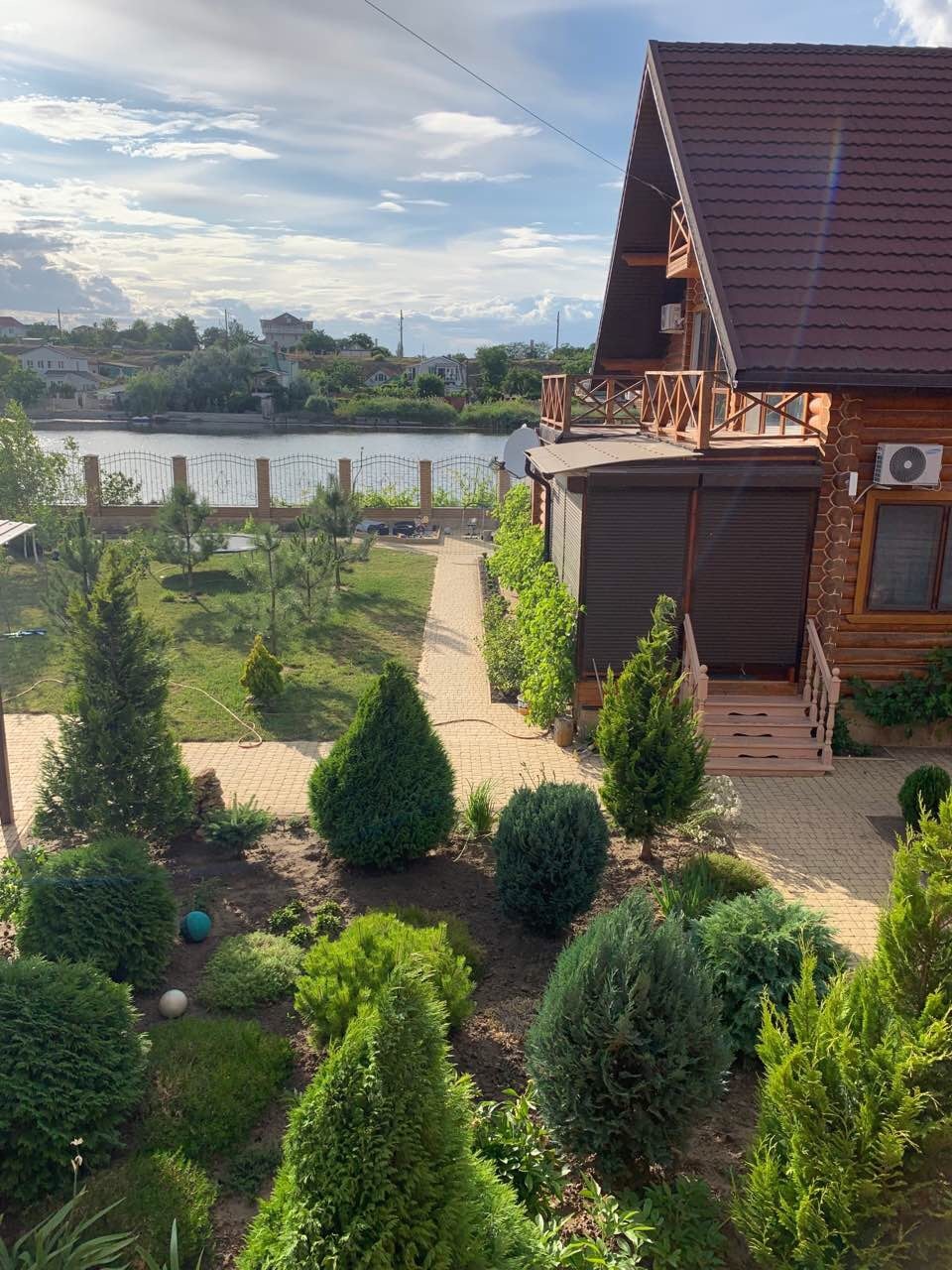 A scenic view of the villa reveals manicured garden areas featuring a variety of lush greenery. A pathway leads towards the lake, which can be seen in the background, with the tranquil water reflecting the sky. The two-story structure is framed by neatly arranged plants.