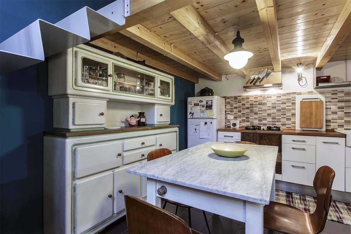 The kitchen features wooden beams and a light wood ceiling. A marble-topped table is surrounded by four wooden chairs. A vintage-style cabinet stands against the wall, displaying glassware. The cabinetry has a modern design with tile backsplash accentuating the cooking area.