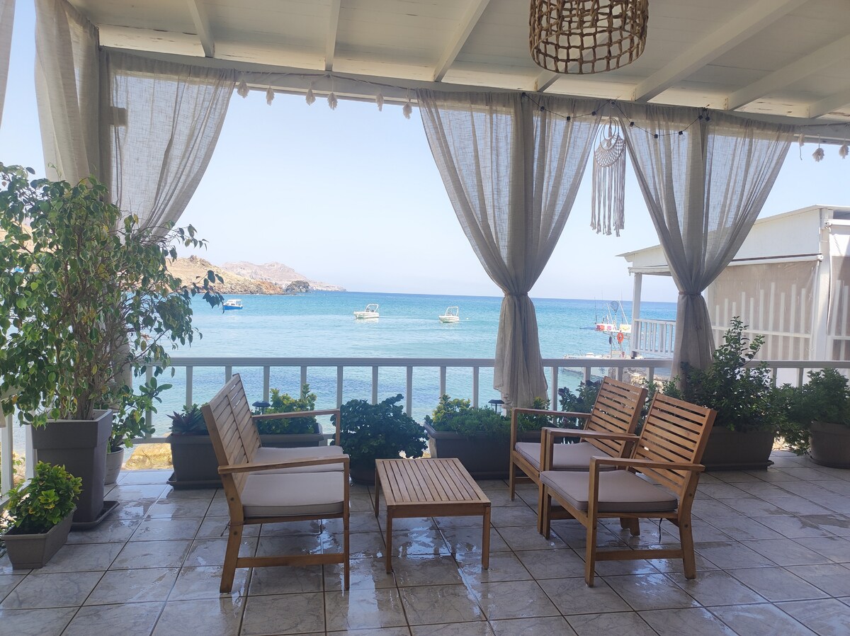A shaded outdoor terrace features wooden seating with white cushions and a low table. Potted plants adorn the area, offering greenery against the tiled floor. The sea and boats are visible in the background, framed by light curtains swaying in the breeze.