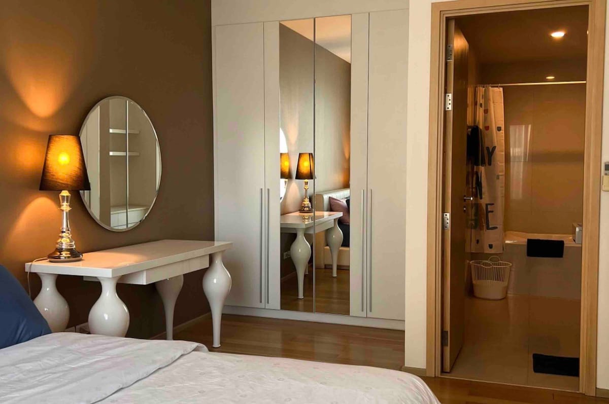 A contemporary bedroom is shown, featuring a spacious bed with neutral bedding. A circular mirror and a stylish table are visible in the foreground. The mirrored wardrobe reflects light and creates depth. A bathroom entrance is seen on the right, highlighting a shower and bathtub.