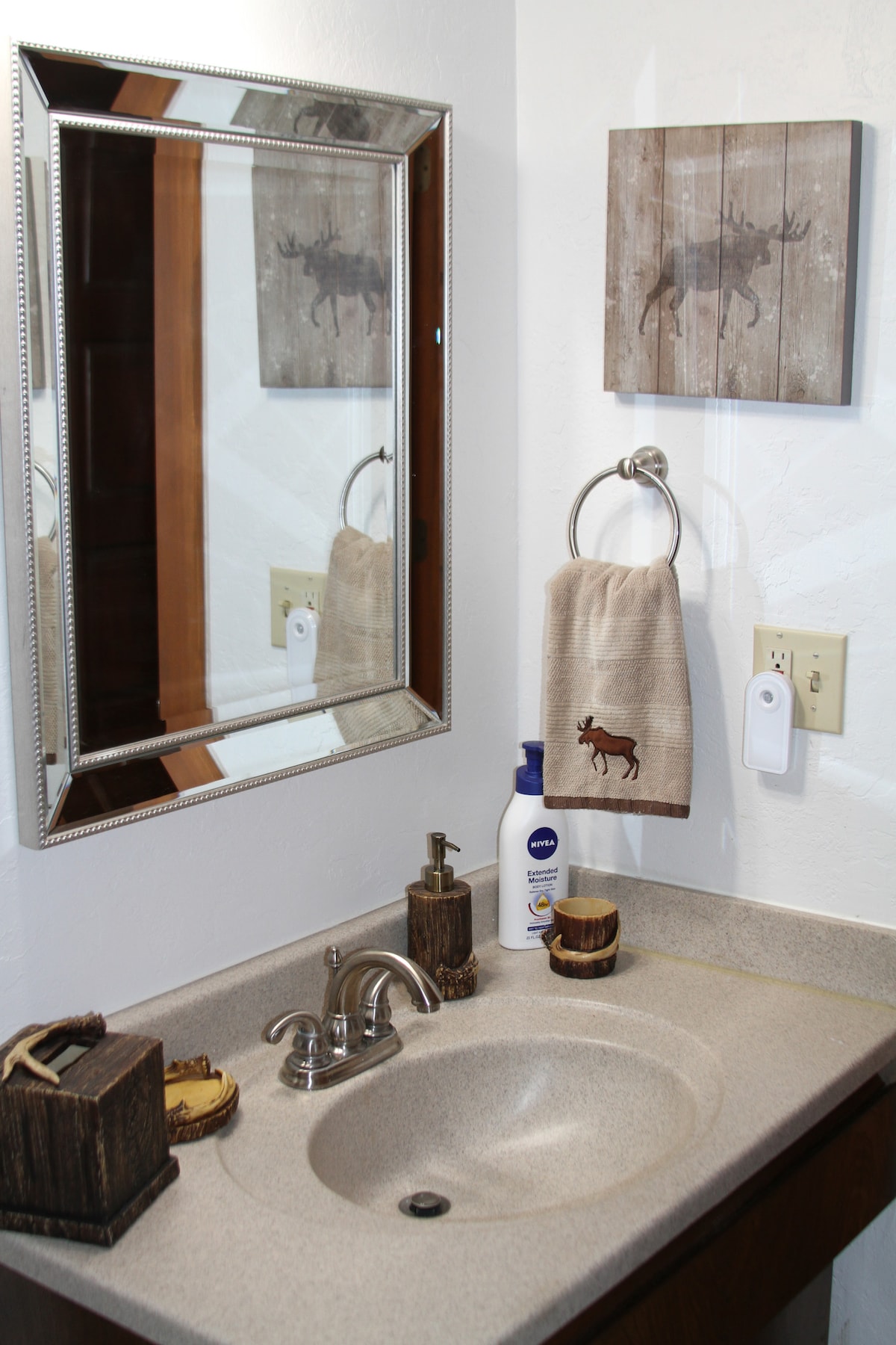 A compact bathroom features a single sink with a smooth countertop. A large mirror reflects the rustic decor, including a wooden moose artwork. A towel, hung on a ring, is accompanied by toiletries neatly arranged on the counter, enhancing the functional space.