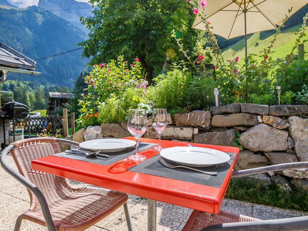 A vibrant red outdoor dining table is set for two, with plates and glasses placed neatly. Lush greenery and colorful flowers surround the area, while a large umbrella provides shade. Majestic mountains are visible in the background, creating a serene atmosphere.