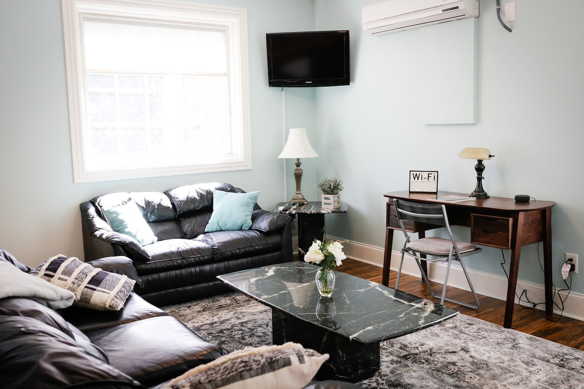 A well-lit living area features a dark leather sofa adorned with soft pillows and a decorative table with a floral arrangement. A wooden desk equipped with a chair supports a Wi-Fi sign, while a flat-screen television is mounted on the wall.