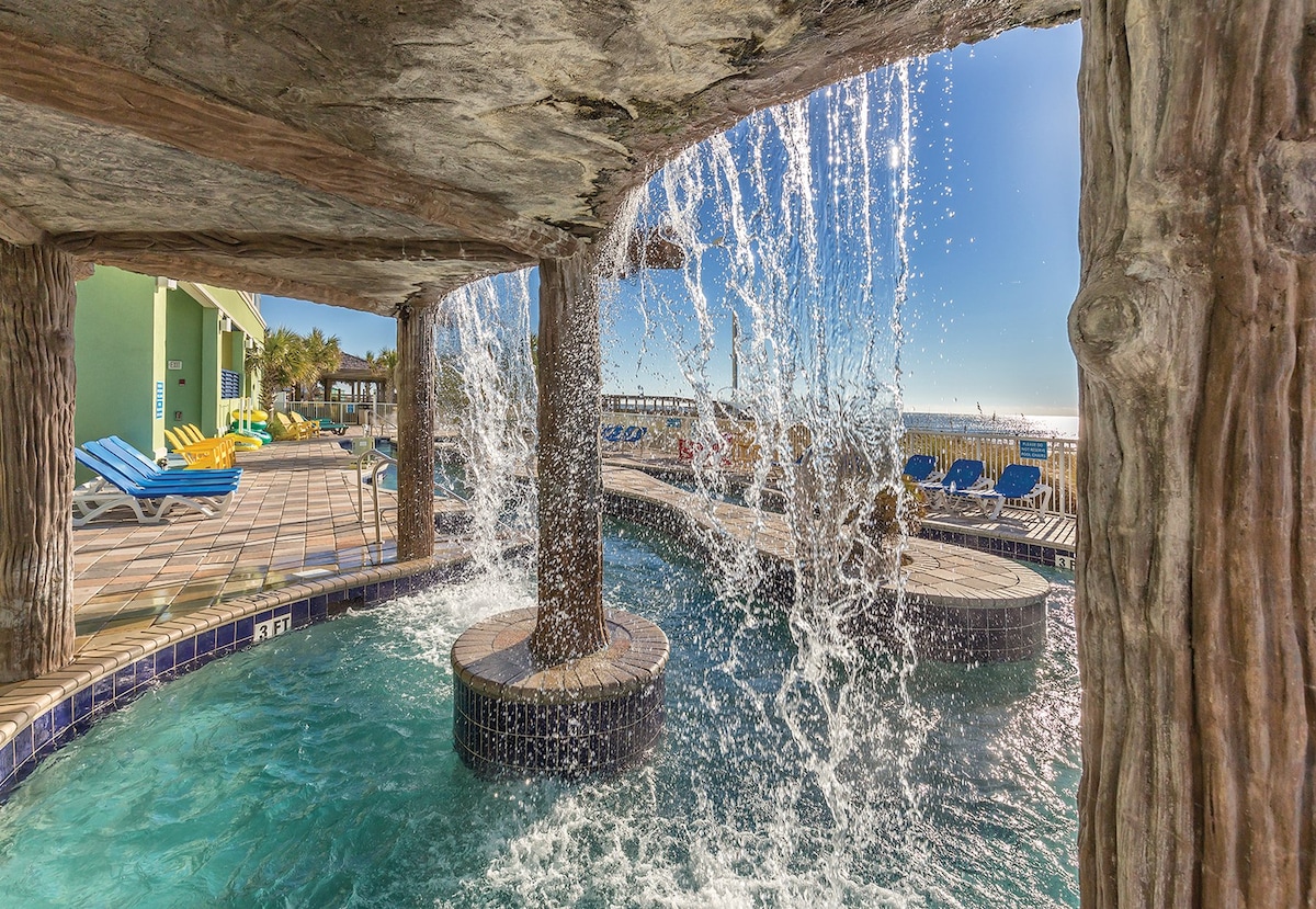 A cascading waterfall flows over natural stone structures, creating a serene atmosphere in a vibrant pool area. Sun loungers can be seen in the background, set against a clear blue sky and the distant ocean view.