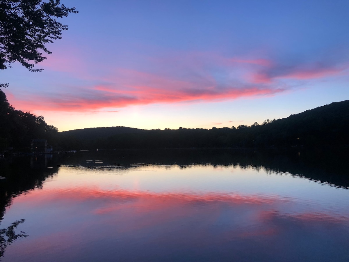 A tranquil lakeside scene at sunset features vibrant pink and orange hues reflected on the calm water. Silhouetted trees line the shore, and rolling mountains rise in the distance, creating a serene backdrop.