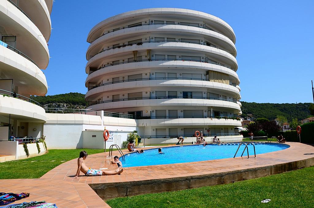 A circular building is prominently featured near a shimmering pool area. The pool is surrounded by a sunlit patio with lounging individuals. Lush green grass borders the pool, creating a relaxing outdoor space. Mountainous terrain can be seen in the background under a clear blue sky.