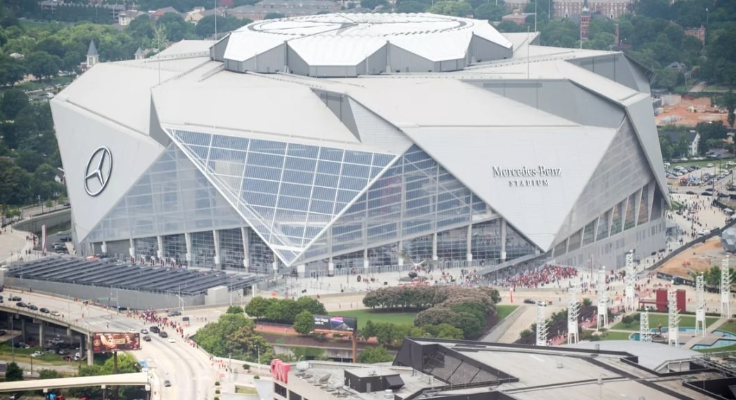An aerial view of the Mercedes-Benz Stadium is presented, showcasing its distinctive exterior design with a large, angular glass facade. Nearby roads and pathways are visible, along with crowds of people approaching the entrance, set against a backdrop of greenery and urban development.