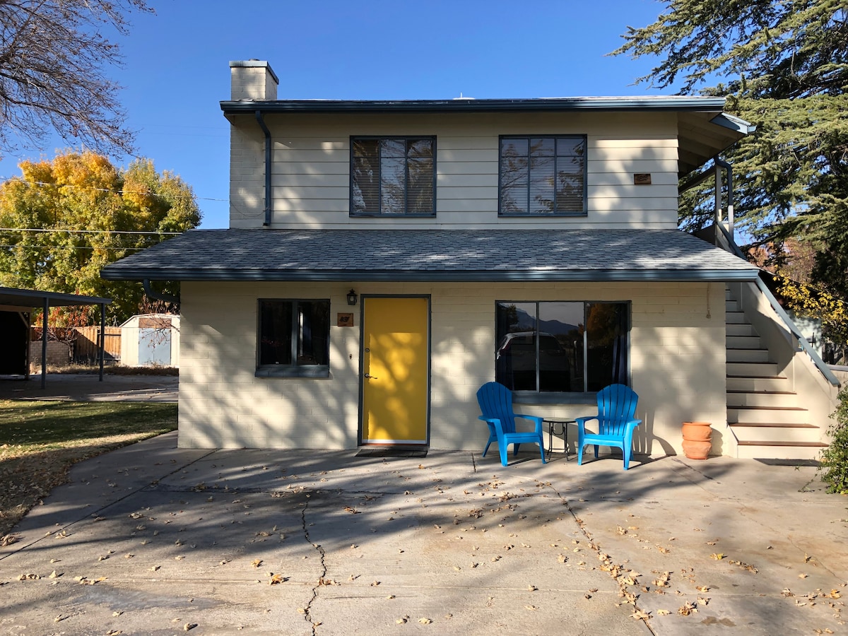 A ground-floor apartment is presented with a welcoming yellow door, flanked by two blue chairs and a small table. Sunlight highlights the exterior, showcasing a well-maintained façade with large windows and a staircase leading to the upper level. Surrounding trees offer shade.