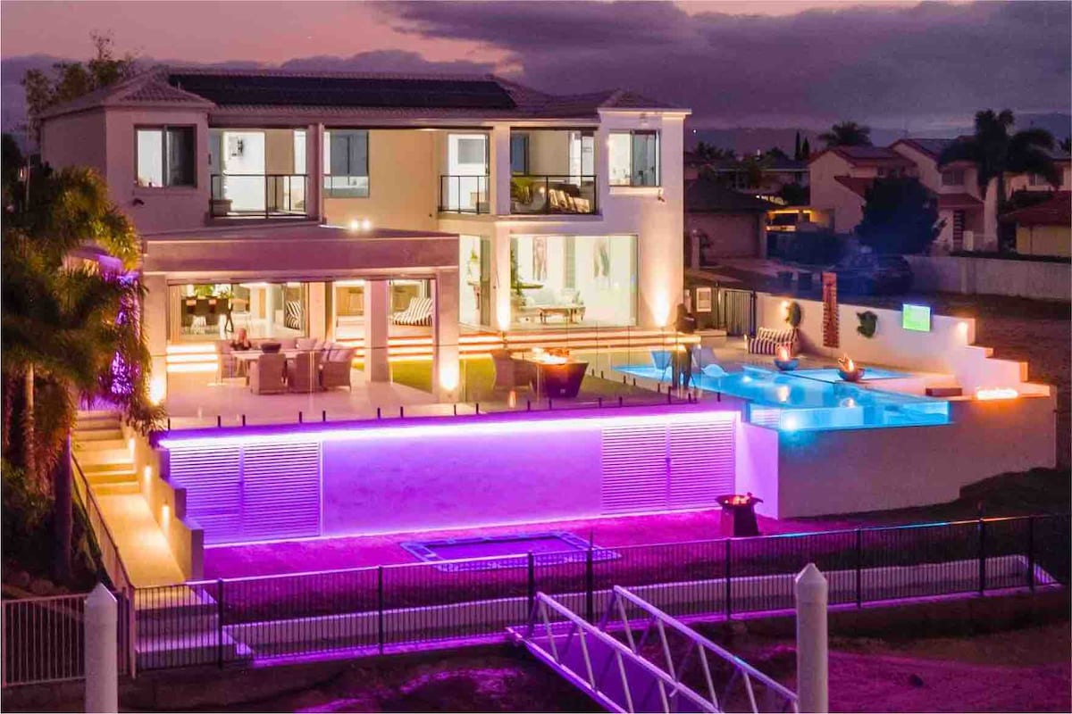 The exterior of a modern waterfront home is illuminated with colorful lighting, featuring a spacious outdoor area with a pool and an alfresco dining space. Multiple levels are visible, and palm trees are set against a twilight sky, creating a vibrant yet serene atmosphere.