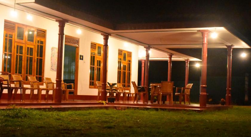 A spacious outdoor dining area is illuminated with soft lighting at night. Multiple wooden chairs surround a table on a covered terrace. The structure features tall windows, allowing views of the garden, while polished columns add an elegant touch.