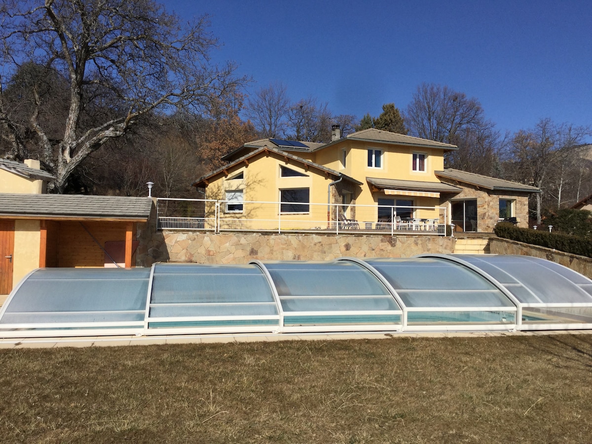 The exterior of a spacious, yellow house is shown against a clear blue sky. A covered pool is positioned in the foreground, with a large terrace visible in the background. A well-maintained garden area surrounds the property, enhancing the serene atmosphere.