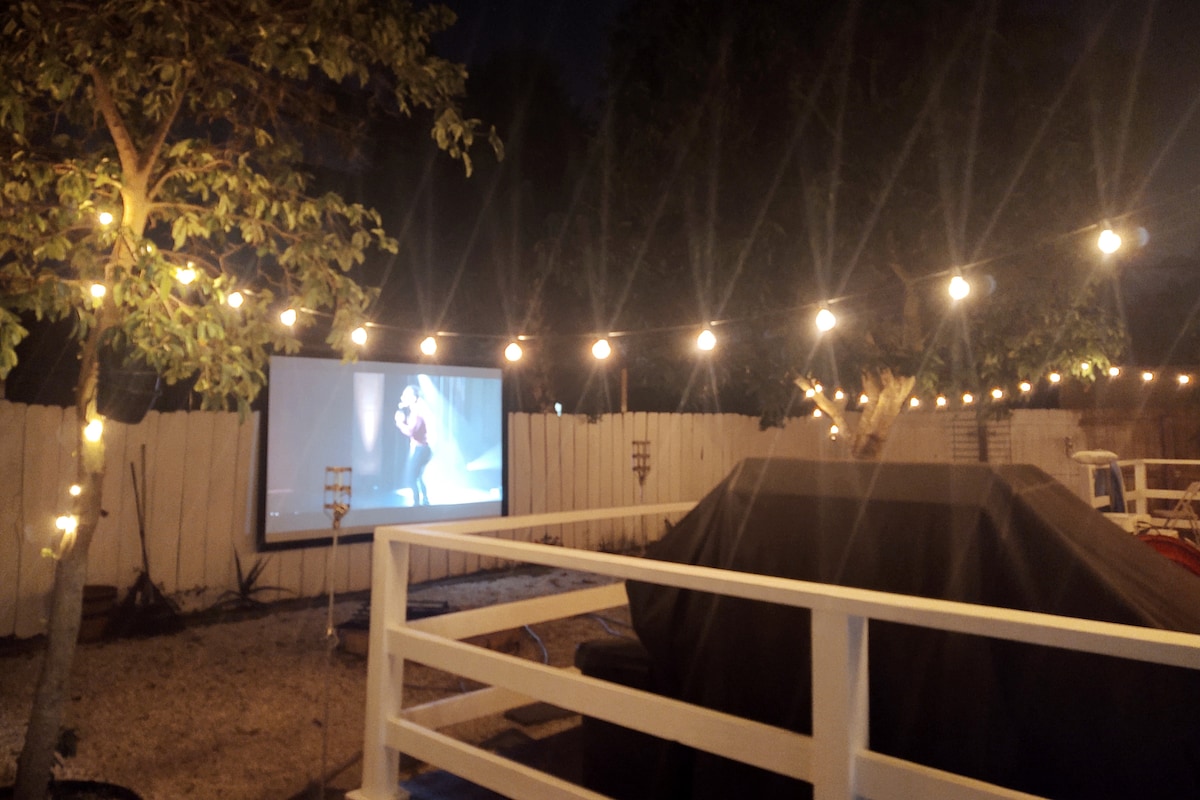 An outdoor space is illuminated by string lights, creating a warm ambiance. A movie screen is set up, showcasing a film. Nearby, a covered BBQ grill is visible, along with a seating area separated by a low white railing and greenery along the perimeter.