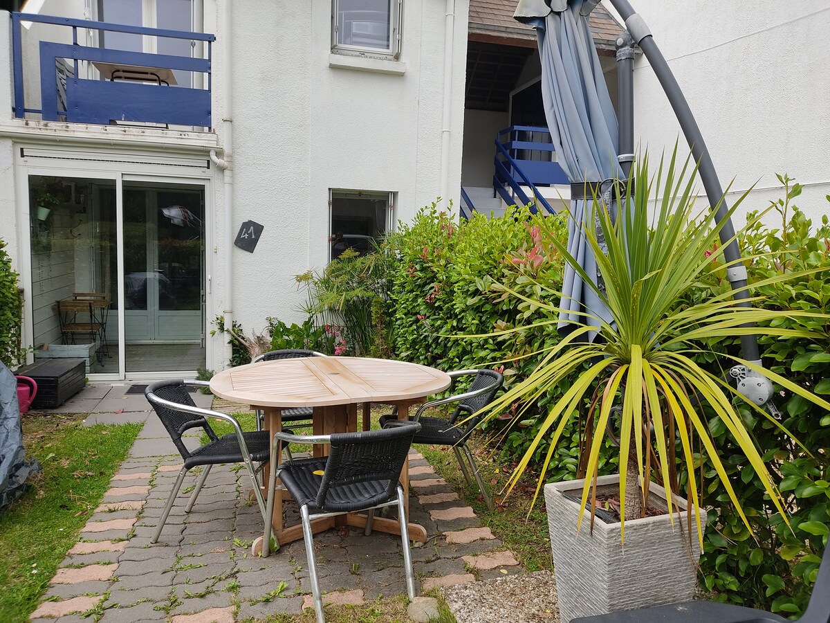 An outdoor dining area is presented with a round wooden table surrounded by black chairs. In the background, a tropical plant adds a splash of greenery. The patio is bordered by neat hedges, and a relaxing seating area is visible to the left.