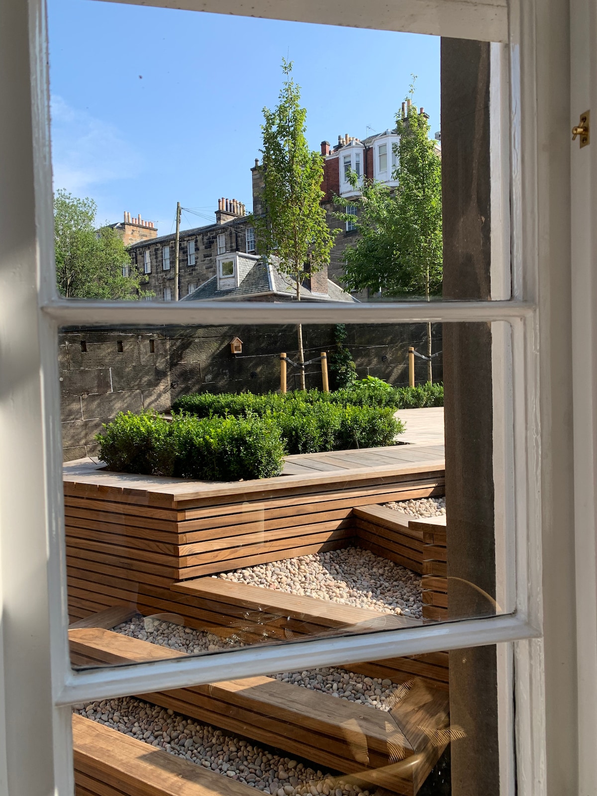 The view through the window frames a landscaped garden featuring tiered wooden decks and pebbled areas. Lush green shrubs complement the natural textures, while tall trees provide shade. The surrounding architecture adds character to the tranquil outdoor space.