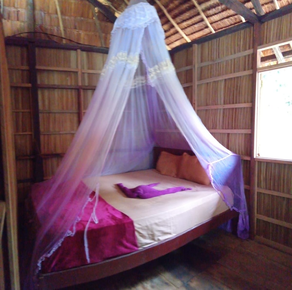 A spacious bed is draped with a delicate mosquito net in soft shades of purple and white. Light filters in through a small window, illuminating the natural wooden walls and ceiling constructed with woven materials.