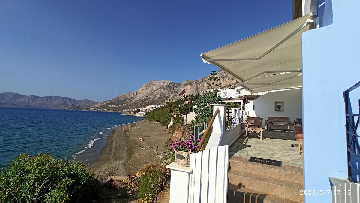 A spacious outdoor terrace overlooks a sandy beach and calm blue waters. The view includes distant mountains lined with residential buildings. Elegant seating is positioned on the terrace, inviting guests to relax and enjoy the scenic coastal landscape.