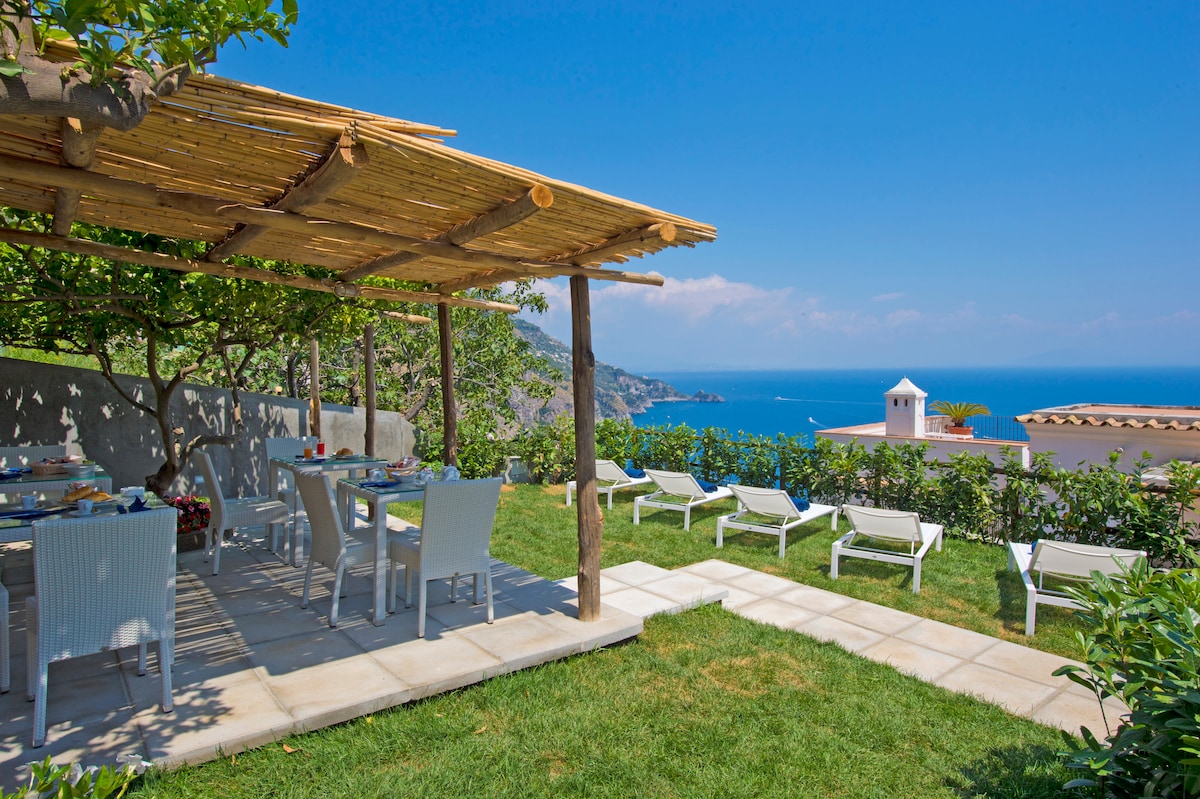 A shaded outdoor dining area features a rustic bamboo pergola, while white tables and chairs are set against a backdrop of lush greenery. Sun loungers are positioned on the grass, and a stunning view of the Mediterranean Sea is visible in the distance.