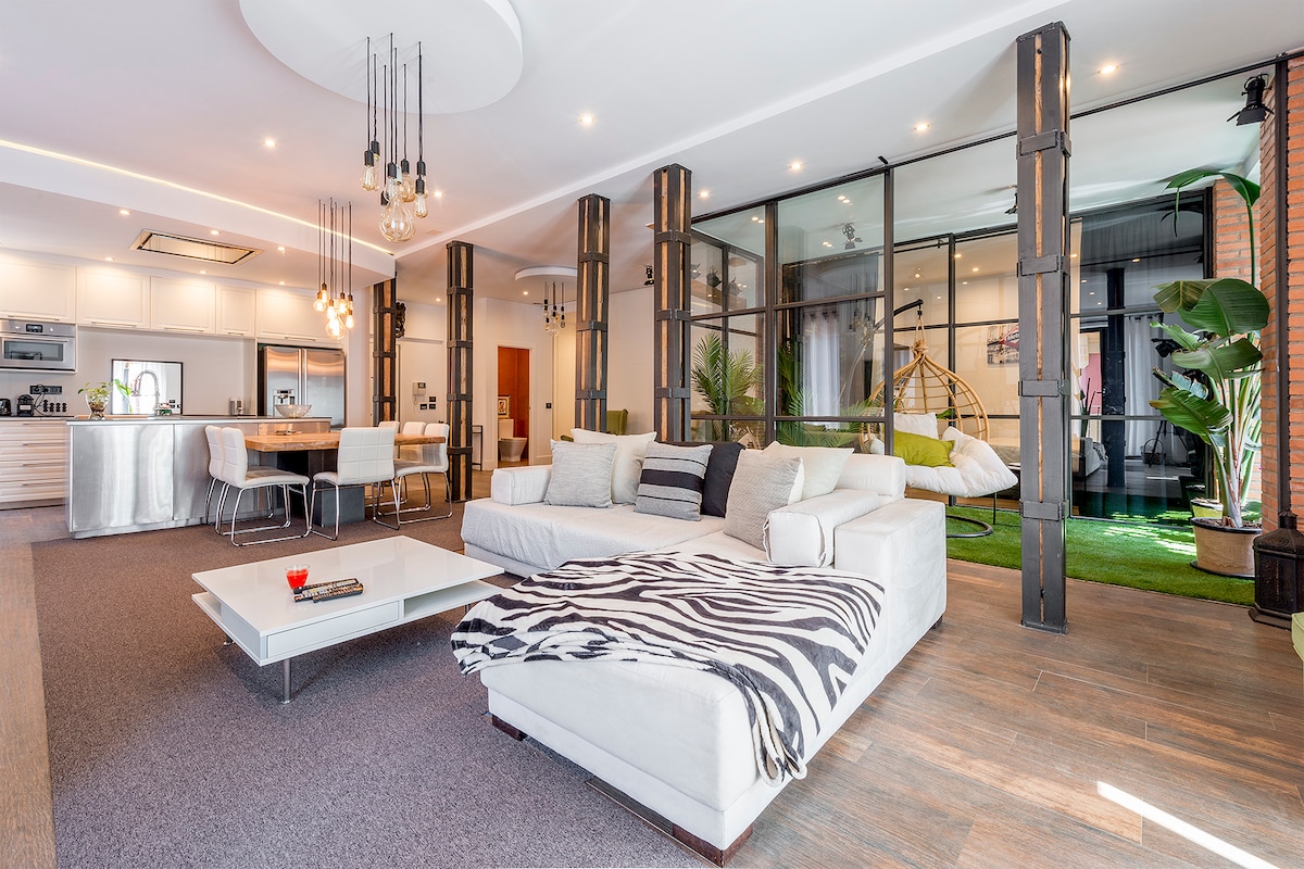 A spacious open-plan living area features a large white sectional sofa adorned with a striped throw. The area is illuminated by pendant lights and framed by large glass panels, with a view of a garden nook and a dining space in the background.