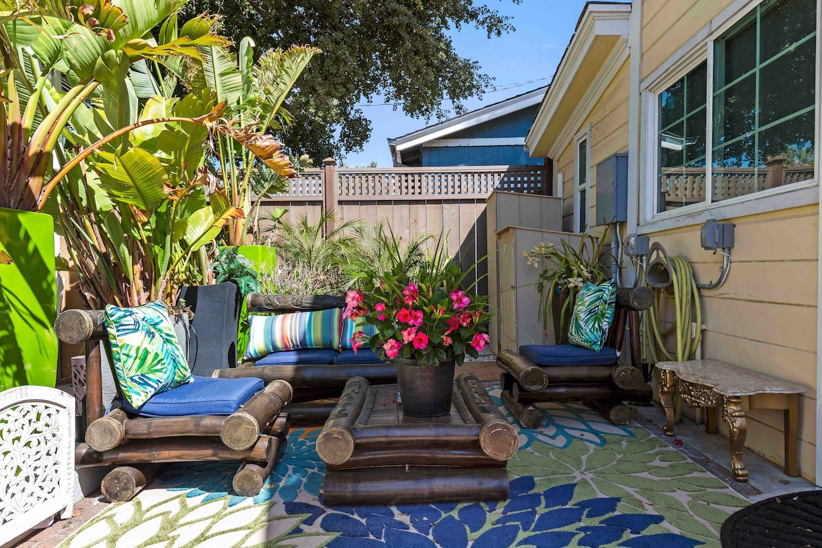 A private patio area is surrounded by lush greenery, featuring comfortable seating with patterned cushions. A colorful floral arrangement is positioned on a wooden table, while a patterned rug adds texture to the outdoor space. Fencing provides privacy and security.