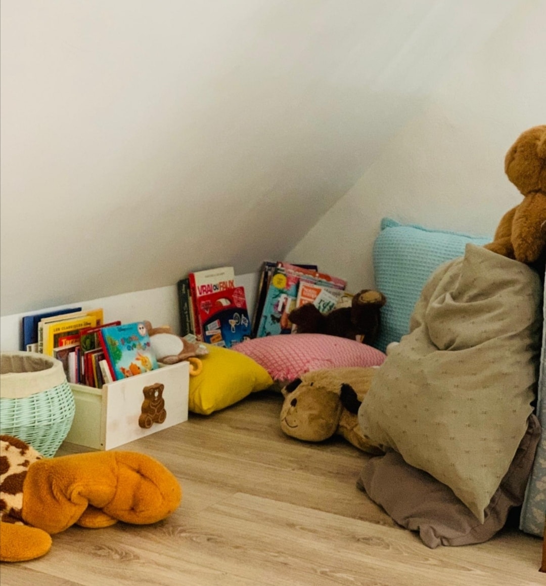 A cozy corner captures a collection of soft cushions in varied colors, providing a comfortable spot for relaxation. Books stacked in a small basket and on the floor suggest a playful reading area. Plush toys are displayed, adding a whimsical touch to the space.