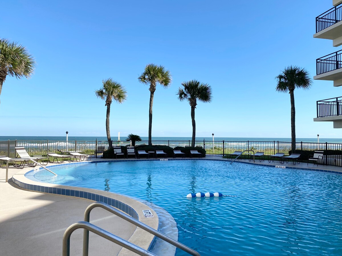A swimming pool is surrounded by palm trees, offering a clear view of the ocean in the background. Lounge chairs are positioned nearby, and the pool area is bathed in natural light from the bright blue sky.