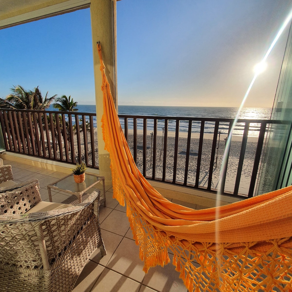 A private balcony with a vibrant orange hammock overlooks the ocean. Comfortable seating is positioned nearby, offering views of the sandy beach and water. Soft sunlight reflects off the waves, enhancing the seaside ambiance.