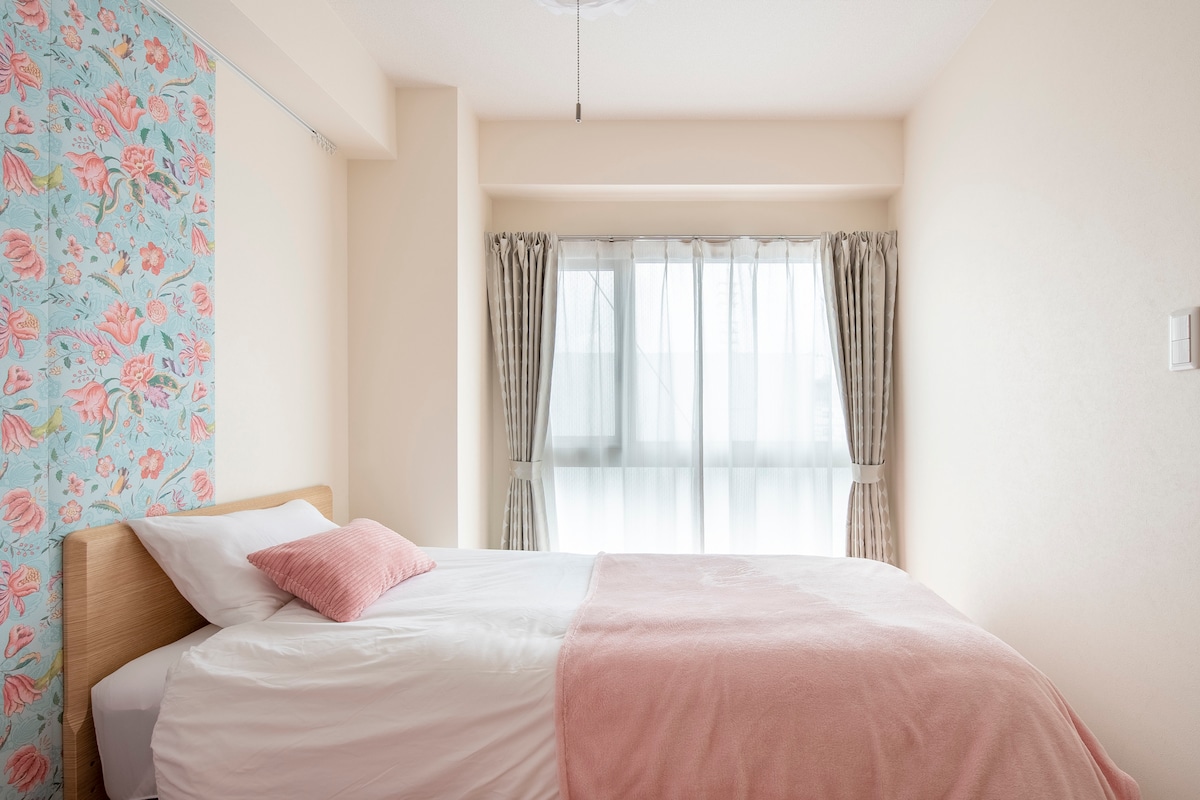 A bright and inviting sleeping area is presented, featuring a spacious bed adorned with a light pink throw blanket. Soft, floral-patterned wallpaper complements the neutral walls. Natural light filters through sheer curtains, illuminating the room while windows provide a view of the outside.