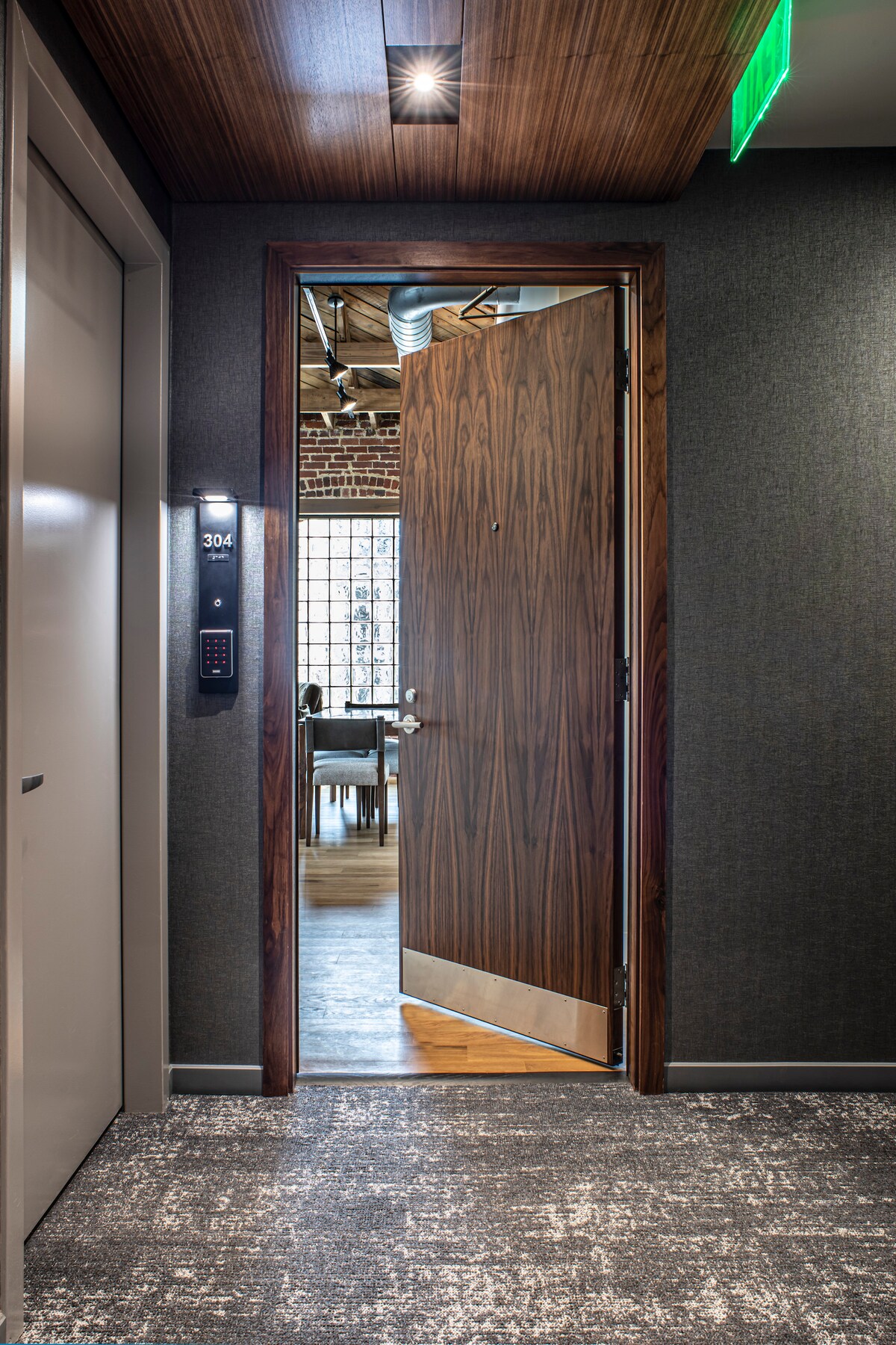 A hallway is shown leading to an open door, revealing a glimpse of a modern interior. The door features a rich wood finish and metal accents, while the textured wall and overhead lighting contribute to a welcoming ambiance.