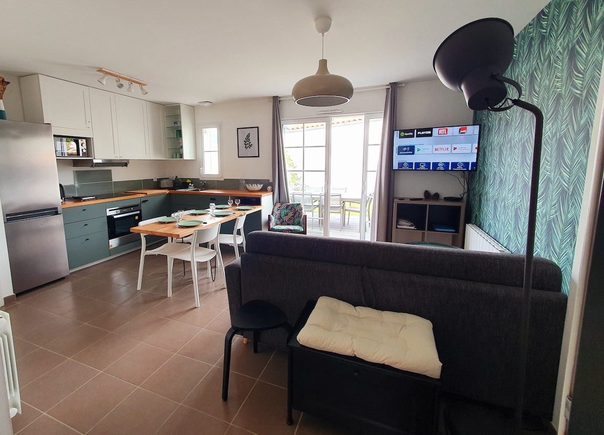 A modern kitchen and living area are shown, featuring a dining table set for four and a comfortable sofa. A connected TV is visible on the wall. Large sliding doors lead to a terrace, allowing natural light to fill the space. Cabinets and appliances are neatly arranged.