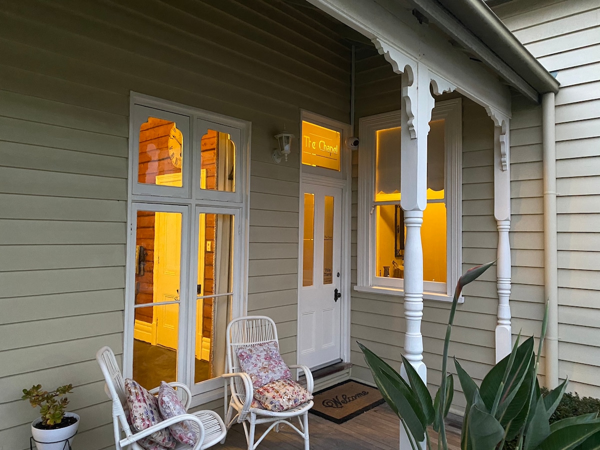 A welcoming porch is presented with two white wicker chairs adorned with floral cushions. A decorative plant stands nearby, and double doors lead to a softly lit interior. The entrance features elegant architectural details, providing an inviting connection to the surrounding environment.