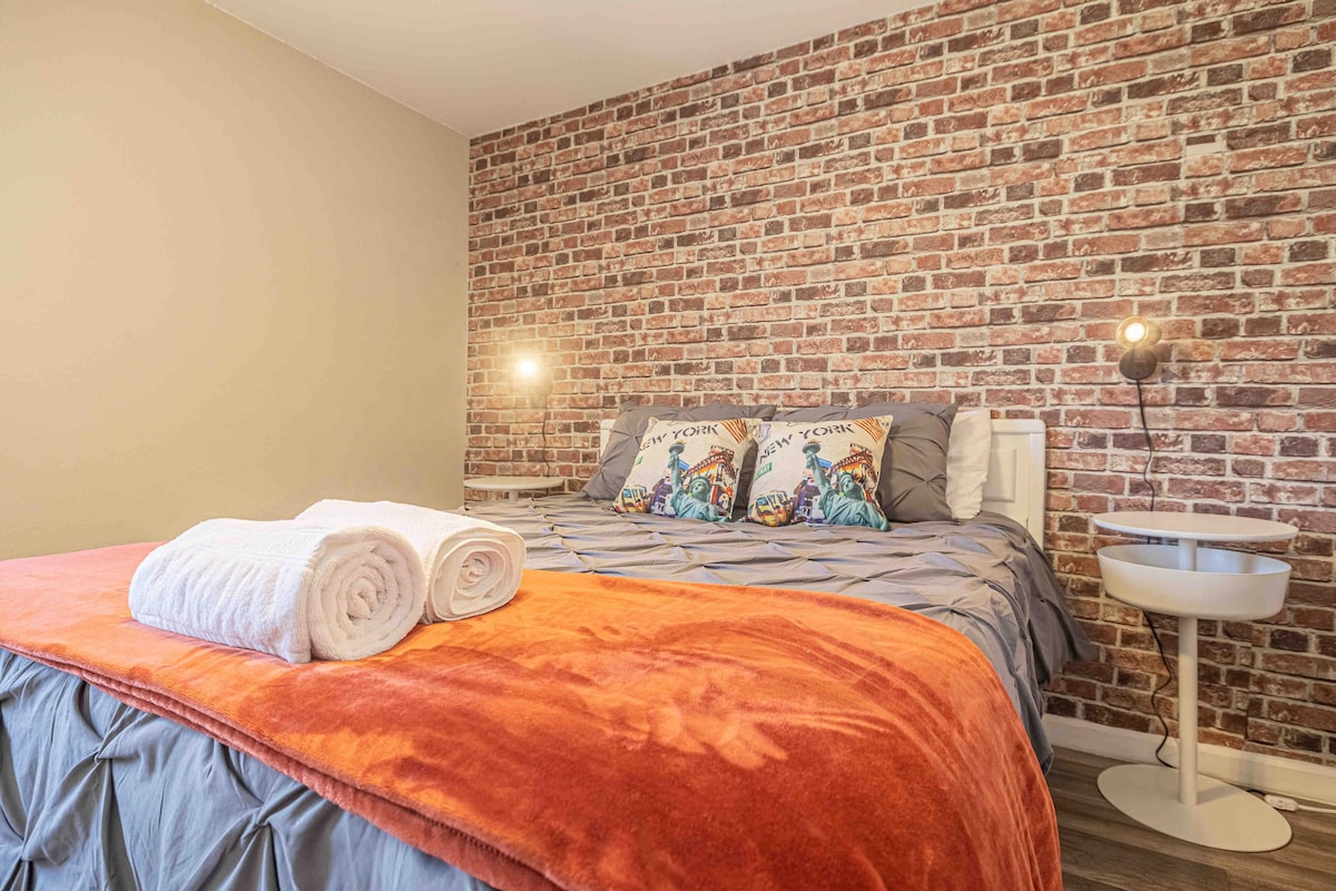 A cozy bedroom features a spacious bed dressed with soft linens and pillows. A warm orange throw blanket drapes over the foot of the bed. The wall is adorned with a textured brick design, and two bedside lamps provide illumination.
