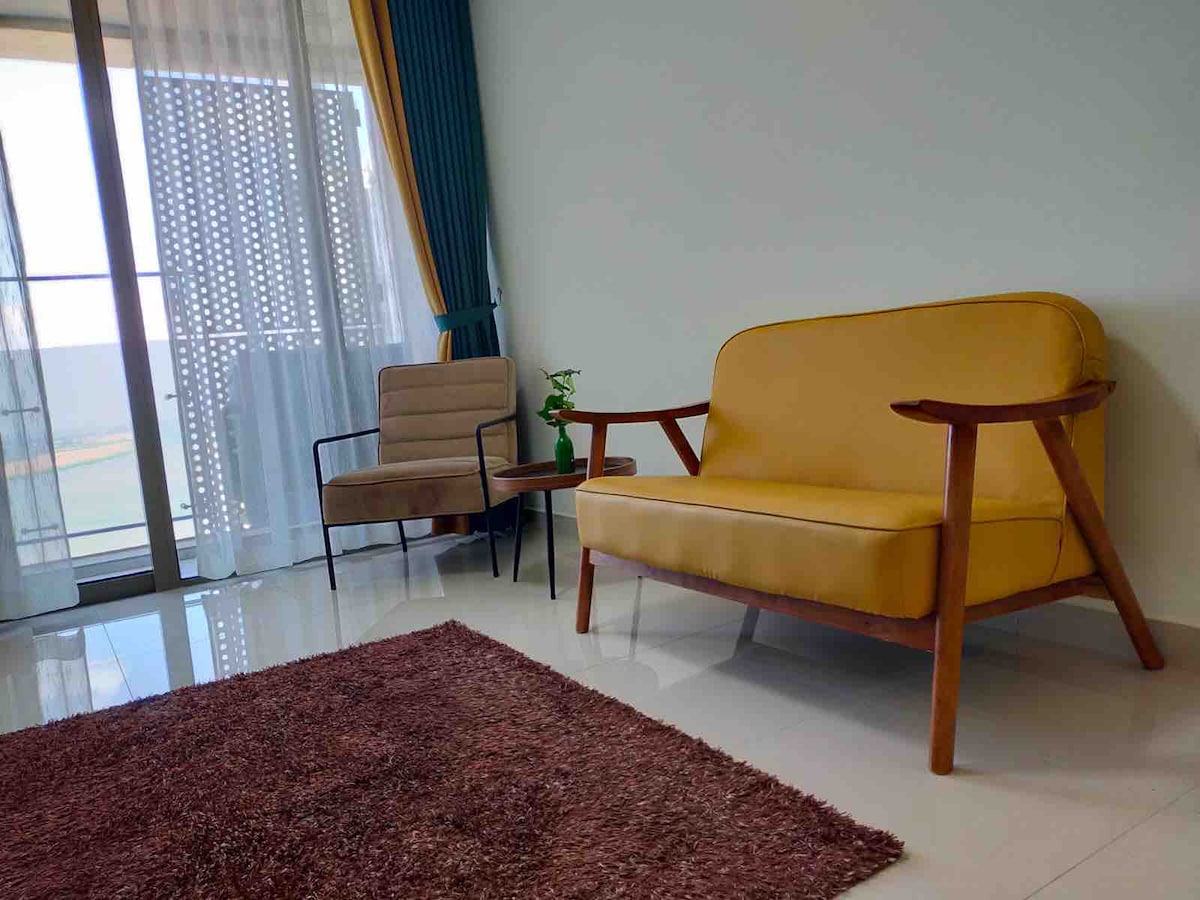 A living area features a yellow sofa and an armchair beside a small table. Natural light filters through sheer curtains, casting soft shadows on a plush, dark rug that enhances the room's comfort. The backdrop includes a large window offering views of the outside.