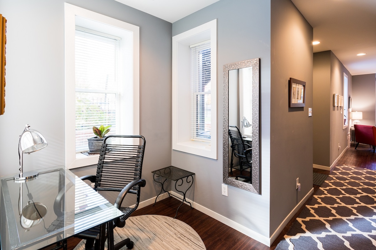 An office area is presented with a glass desk and an adjustable black chair. Natural light enters through a large window, highlighting a small potted plant on the window sill. A wall mirror reflects the space, while a patterned rug adds texture to the floor.