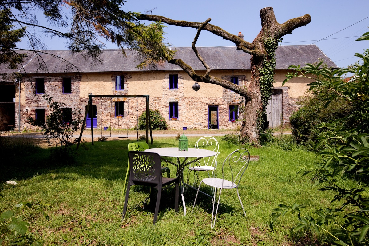 A grassy area features a small table and four chairs set under a tree that has swings attached. The rustic stone building with blue-framed windows is visible in the background. The setting provides a peaceful spot for relaxation and outdoor activities.