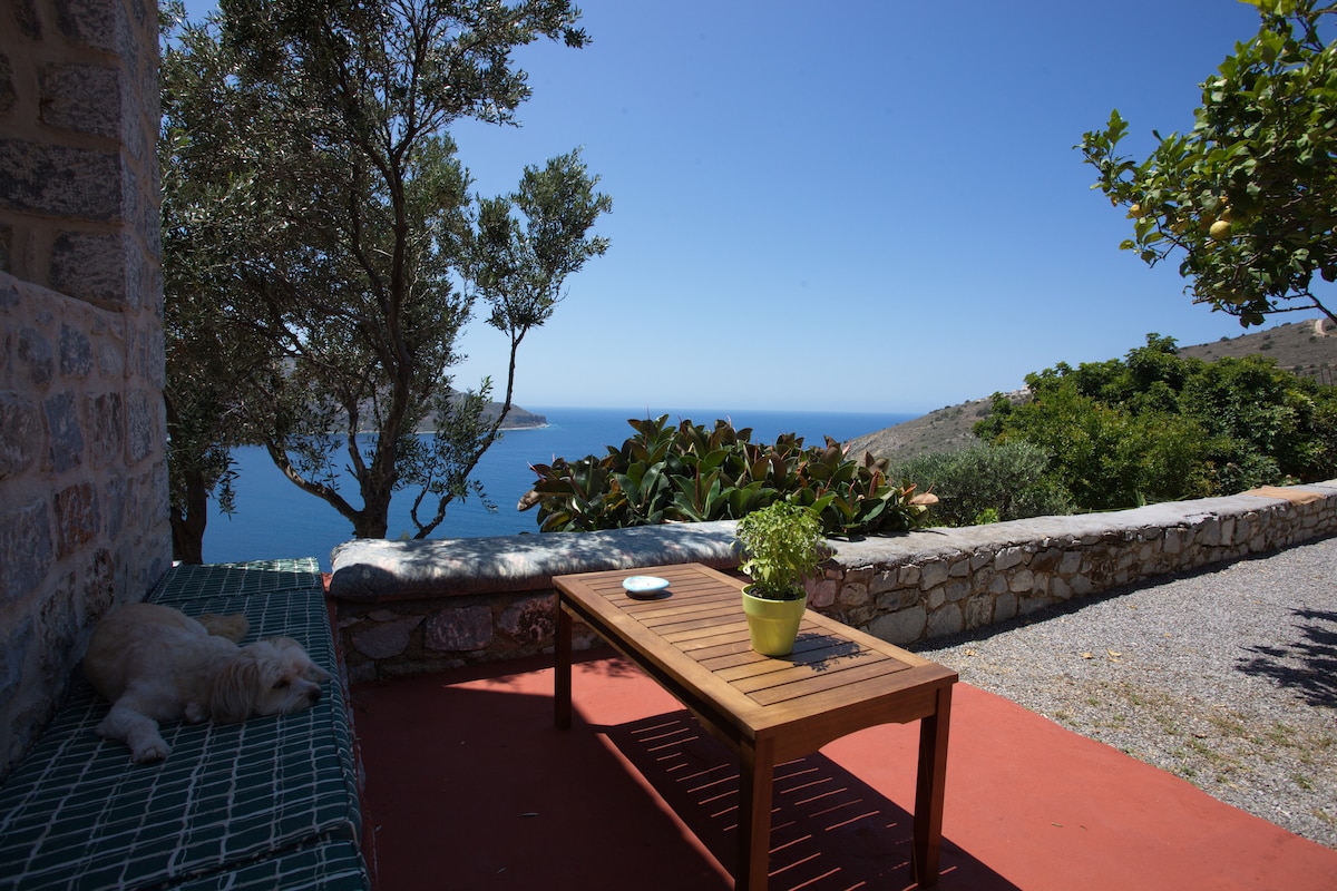 A terrace space features a wooden table with a potted plant at its center, accompanied by a lounge area. The sea and bay are visible in the background, framed by lush greenery and stone walls under a clear blue sky.