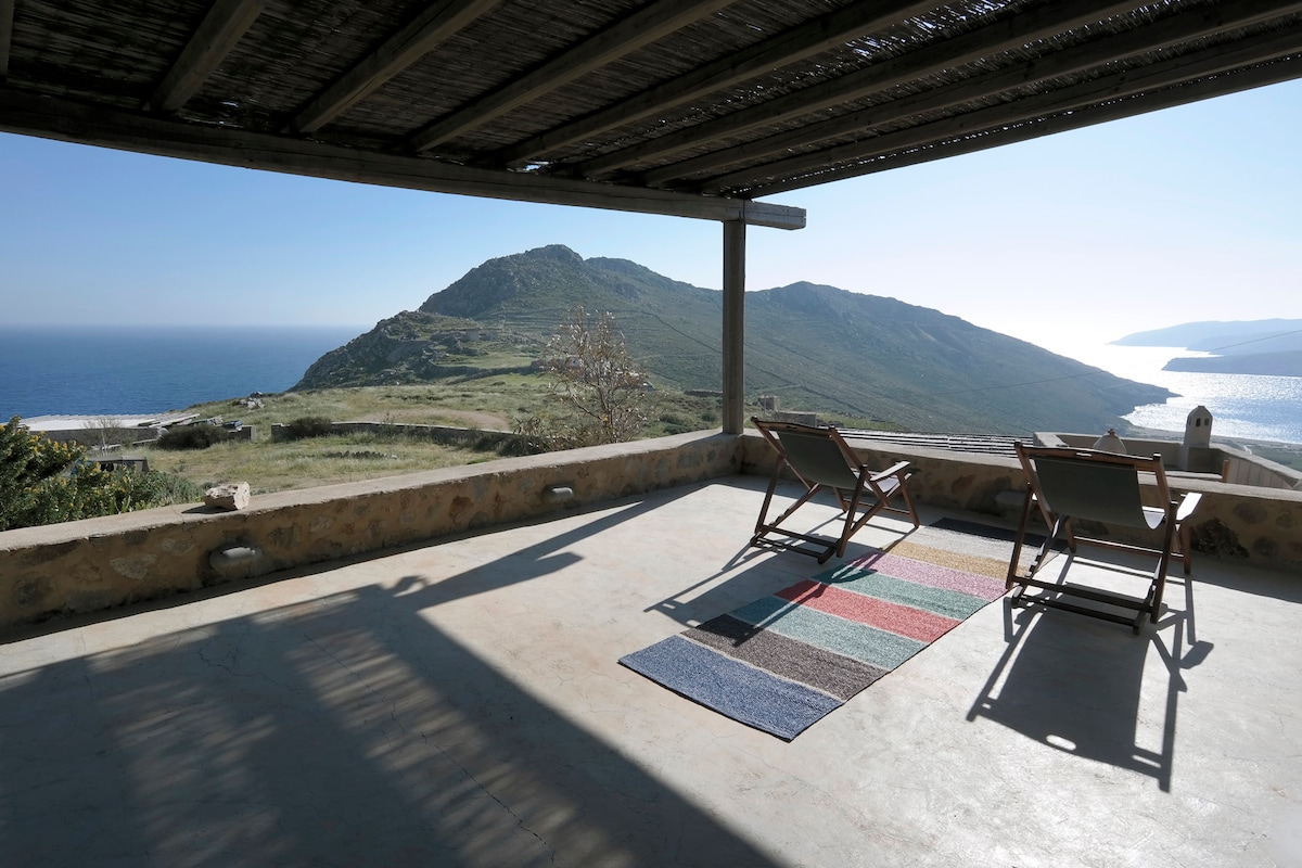 An expansive outdoor terrace is visible, featuring two loungers positioned on a colorful striped rug. The scenic view reveals rolling hills and the coastline along with the serene blue sea under a clear sky.