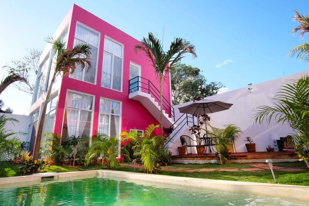 A modern condominium features vibrant pink exterior walls and large windows that allow ample natural light. A lush garden surrounds the property, complemented by palm trees. A shared pool is framed by greenery, with a shaded seating area under an umbrella, creating a relaxed outdoor environment.
