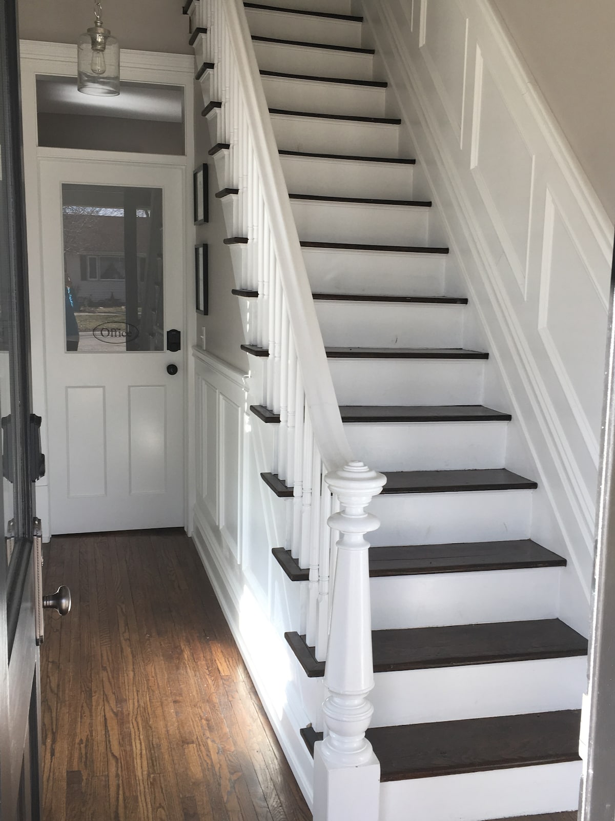 A well-lit staircase is visible, featuring dark wooden steps and a white railing. Natural light streams in from a nearby door with glass panels, illuminating the clean lines of the walls and the polished hardwood floor.