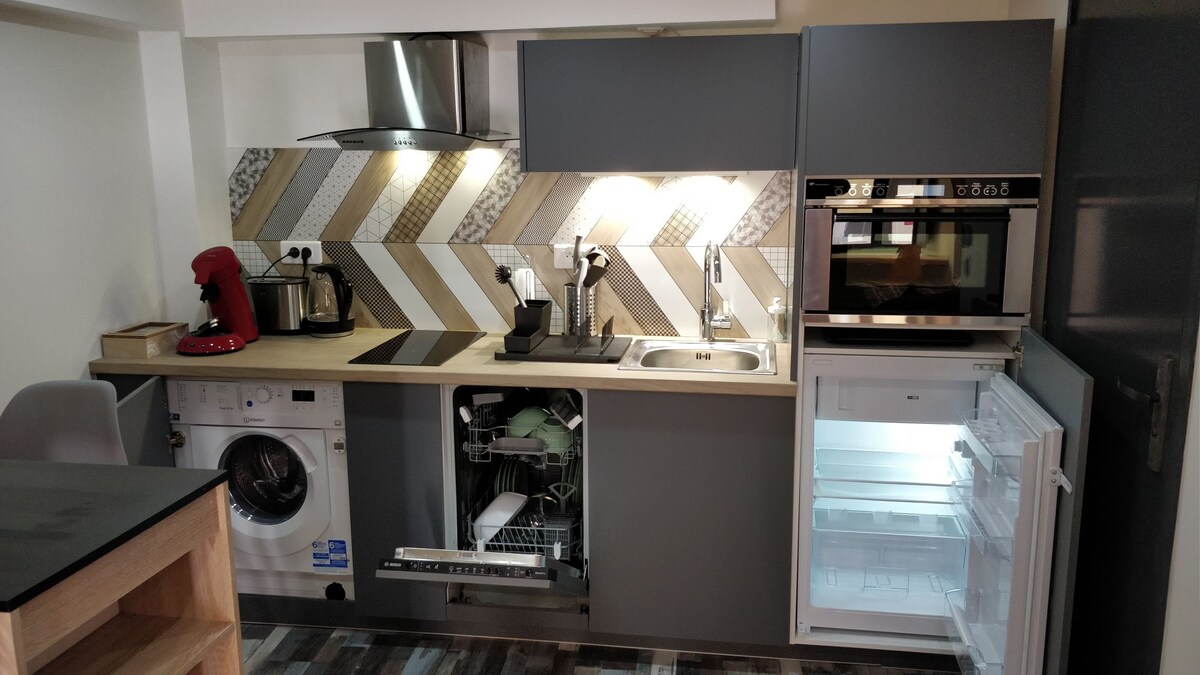 A modern kitchen featuring a variety of appliances, including a washing machine, dishwasher, and multifunction oven. Counter space is complemented by a sleek stove and coffee machine, with a refrigerator and storage cabinets visible, showcasing a functional and well-equipped cooking area.