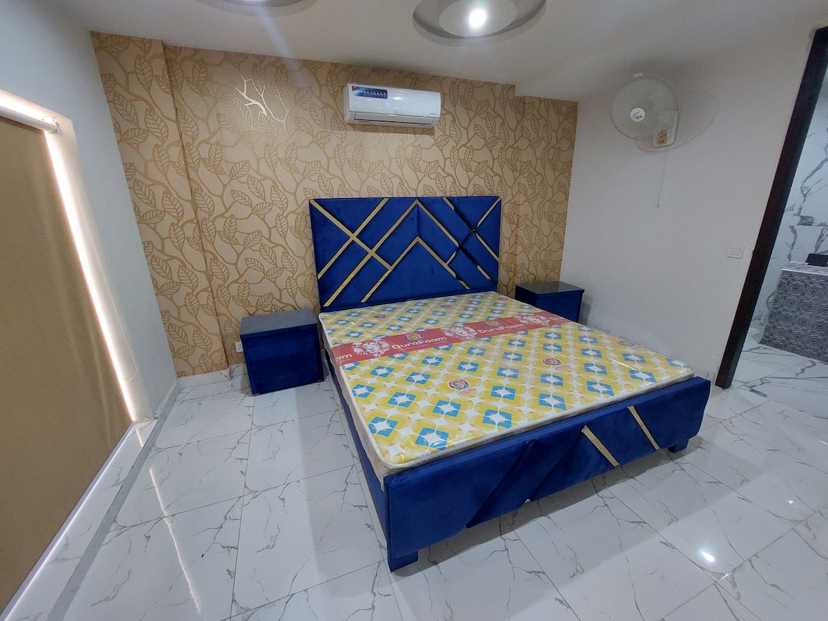 A stylish bedroom features a spacious, blue upholstered bed framed by geometric patterns. Two bedside tables are positioned on either side of the bed, while a wall adorned with a subtle design adds visual interest. Bright lighting enhances the room's contemporary feel.
