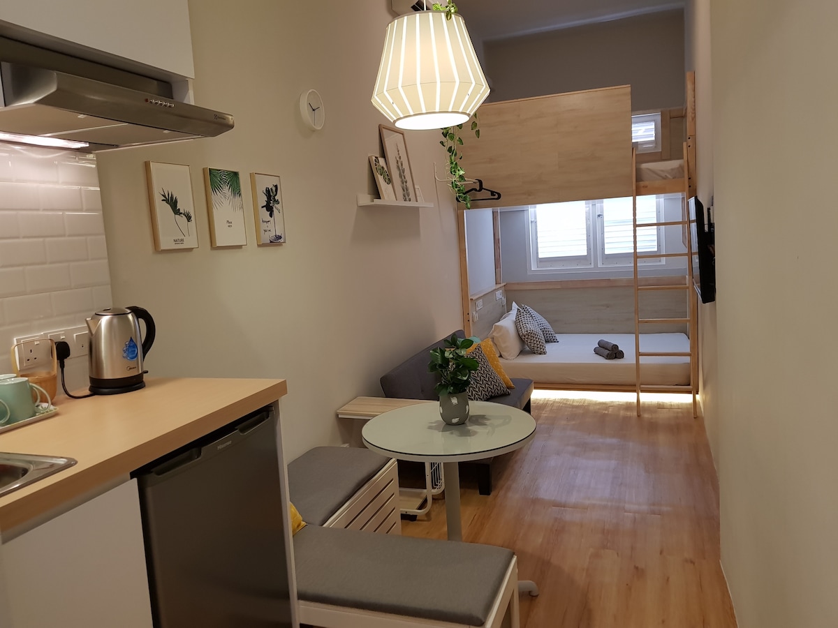 A compact studio layout is visible, featuring a kitchenette with a kettle and a refrigerator. A round table is surrounded by cushion benches. A cozy sleeping area is highlighted by a loft bed and a sofa, offering a calm and functional living space.