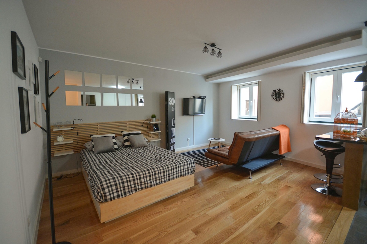 A spacious room features a wooden bed with checkered bedding, complemented by two grey pillows. A modern sofa bed is positioned nearby, alongside a small bar table with stools. Large windows allow natural light, enhancing the warm wooden flooring and minimalist decor.