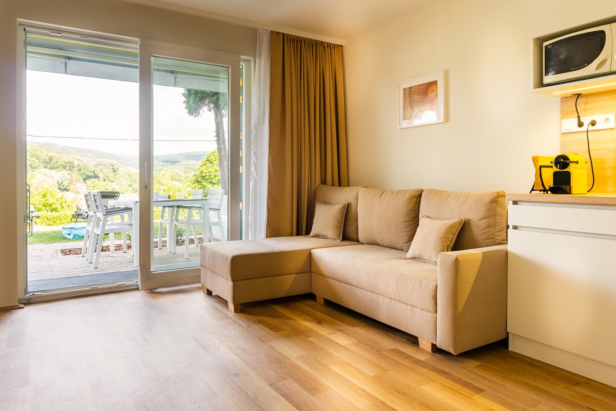 A spacious living area features a light-colored, L-shaped sofa beside large glass doors that open to a scenic view of the outdoors. Natural light fills the room, complemented by wooden flooring. A compact kitchen area is also visible, providing essential amenities.